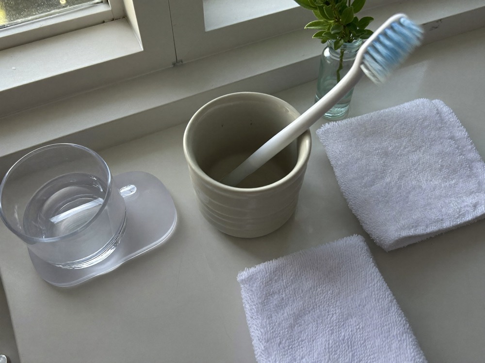 A toothbrush in a ceramic cup, a glass of water and two folded white washcloths on a bathroom counter by a window.