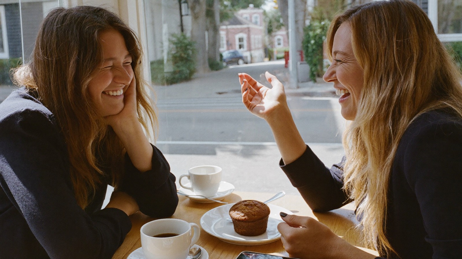 Two women laughing over coffee at a sunny café window table.