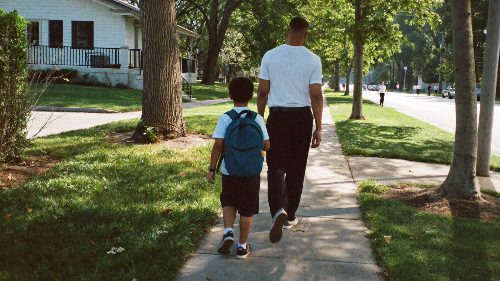 A man and a young boy with a blue backpack walking along a tree-lined sidewalk past front porches on a sunny morning.