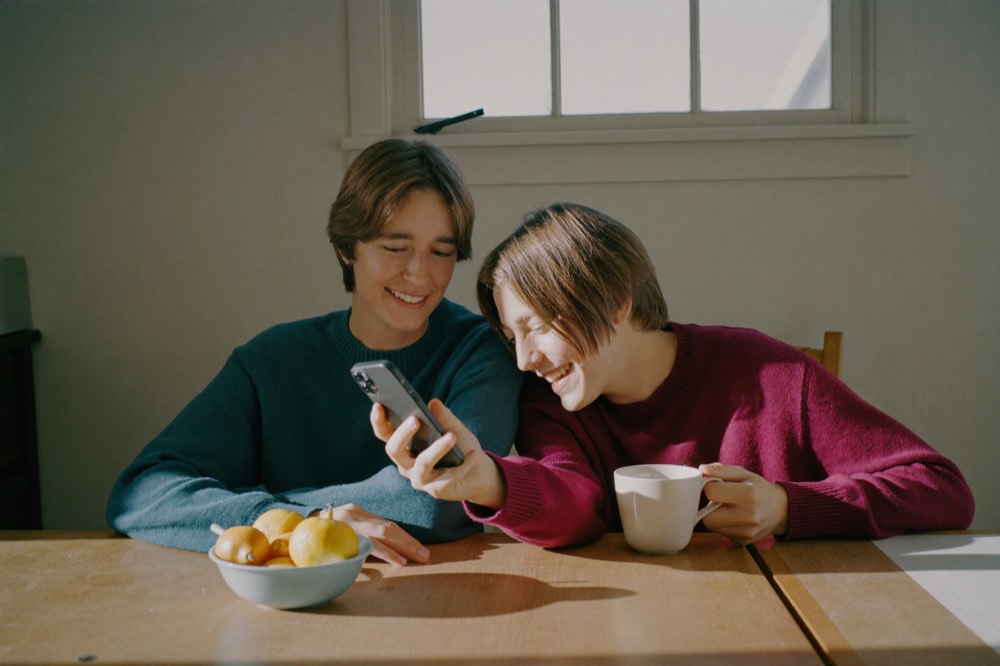 Two young people in sweaters laughing as they look at a phone together at a sunny kitchen table, with a mug and a bowl of fruit.